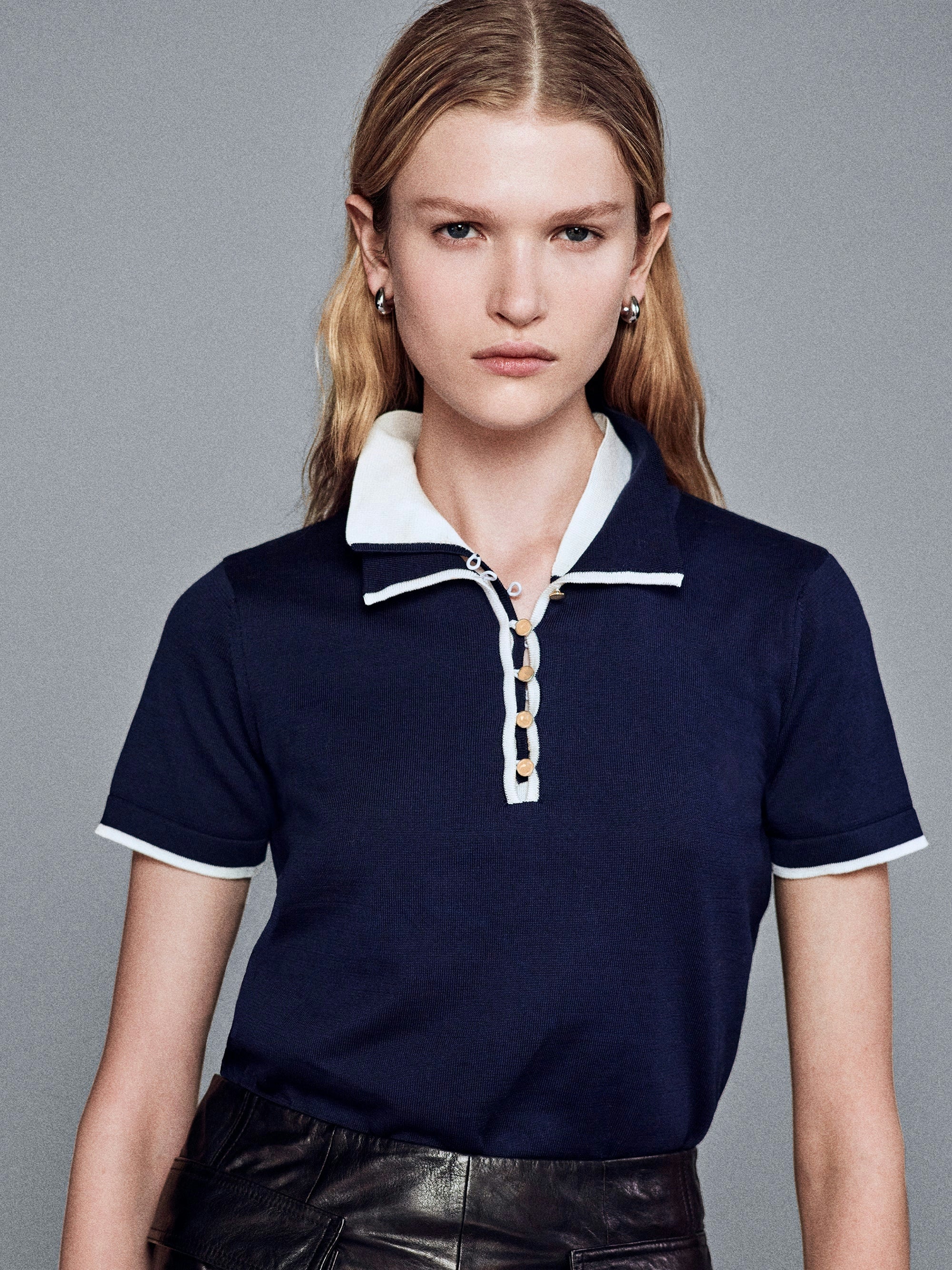 A woman wearing a navy blue polo shirt with white contrast trim at the inner collar, button placket, and short sleeve hem, and gold dome buttons.