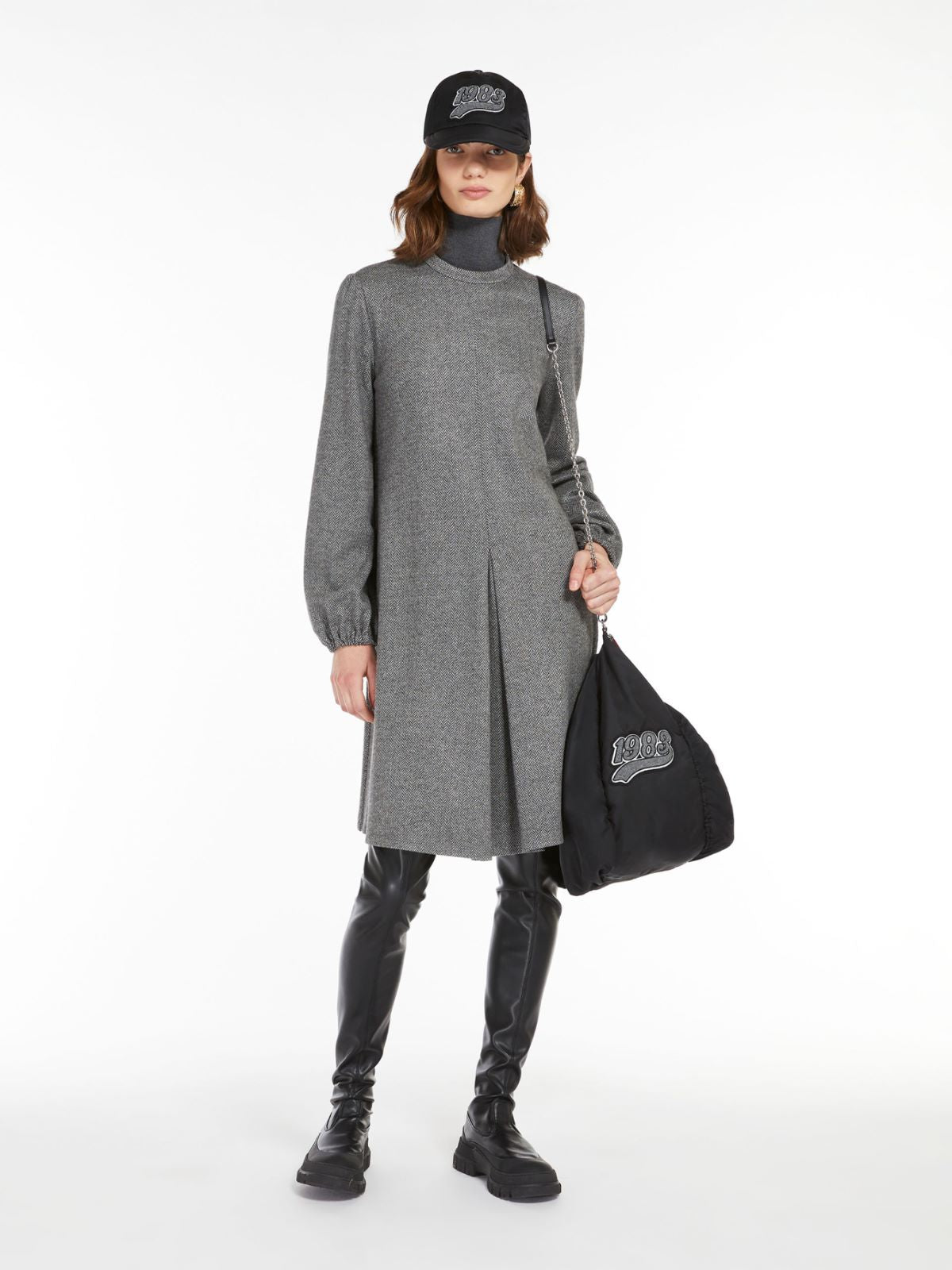 A model wearing a long sleeve, high neck, gray dress with an inverted pleat in the front, paired with black leggings and carrying a black bag with a logo on it.