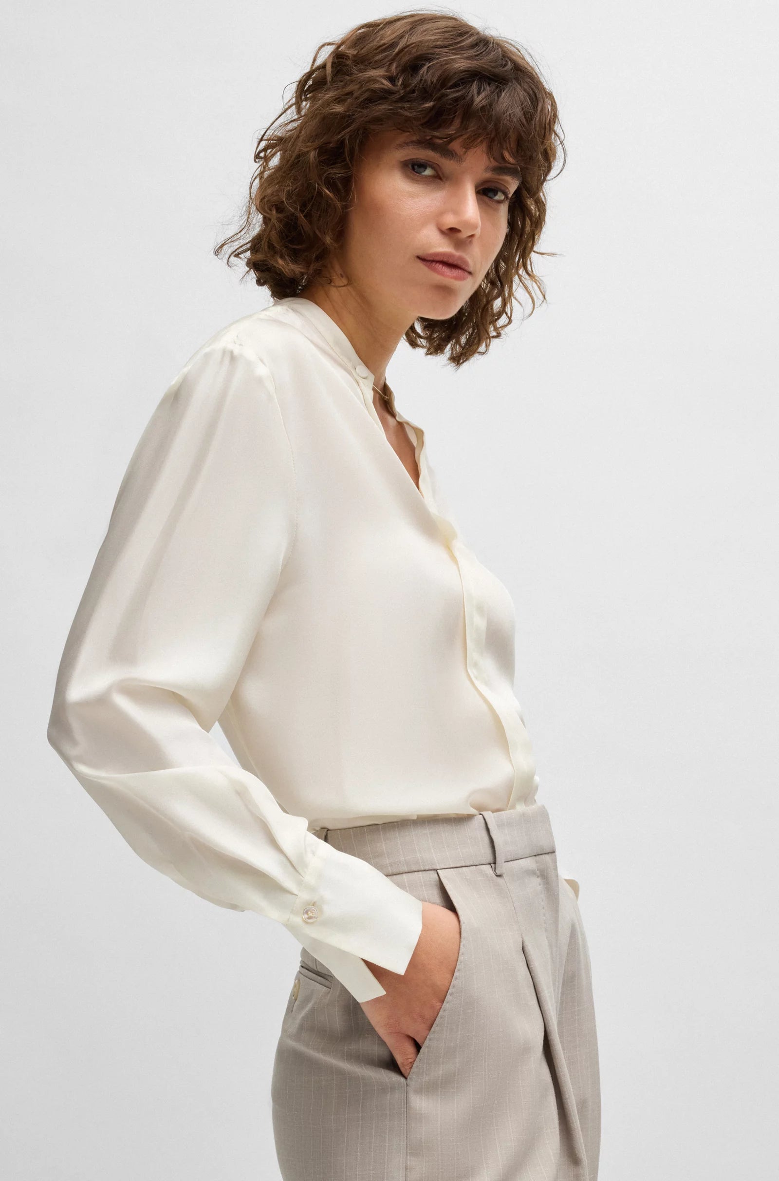 A woman wearing a beige, relaxed-fit blouse with long sleeves and a stand collar. The blouse is made of Italian silk and has a drawstring closure at the neck.