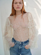 A woman wearing a beige ruffle blouse with a V-neckline and long sleeves, paired with blue jeans.