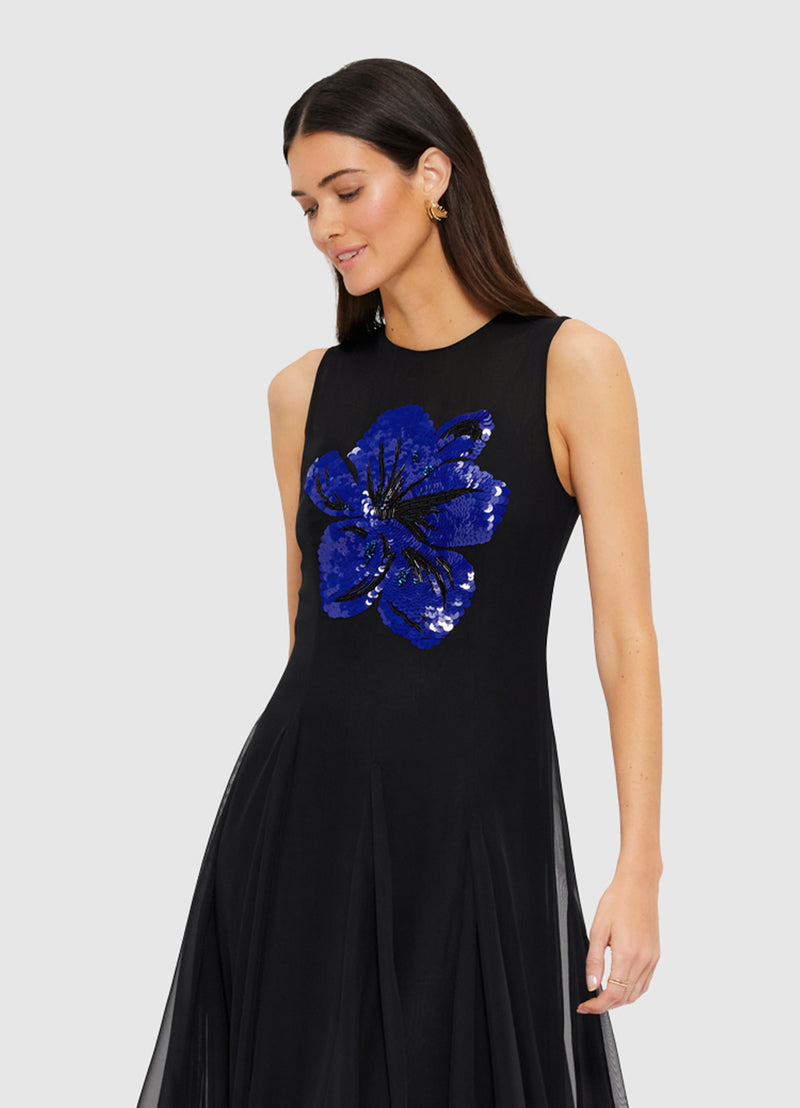 A woman wearing a black maxi dress with a round neckline and embellished floral motif, with a fitted waistline and A-line maxi skirt.