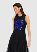 A woman wearing a black maxi dress with a round neckline and embellished floral motif, with a fitted waistline and A-line maxi skirt.