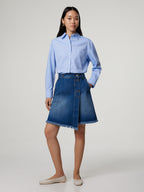 A person standing in a studio wearing a blue denim skirt with an asymmetric fringe hem and back patch pockets, paired with a blue shirt.