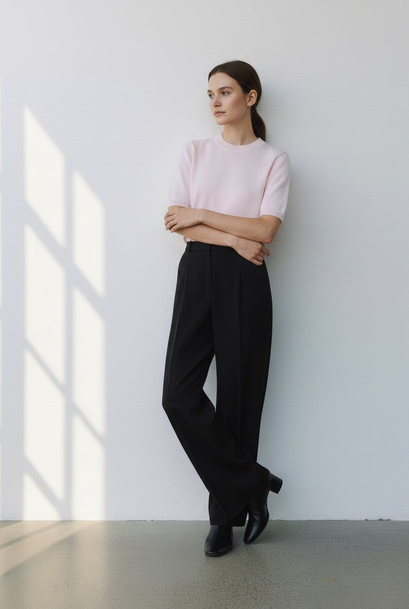 Person wearing a light pink top and black pants standing against a white wall with sunlight streaming in.