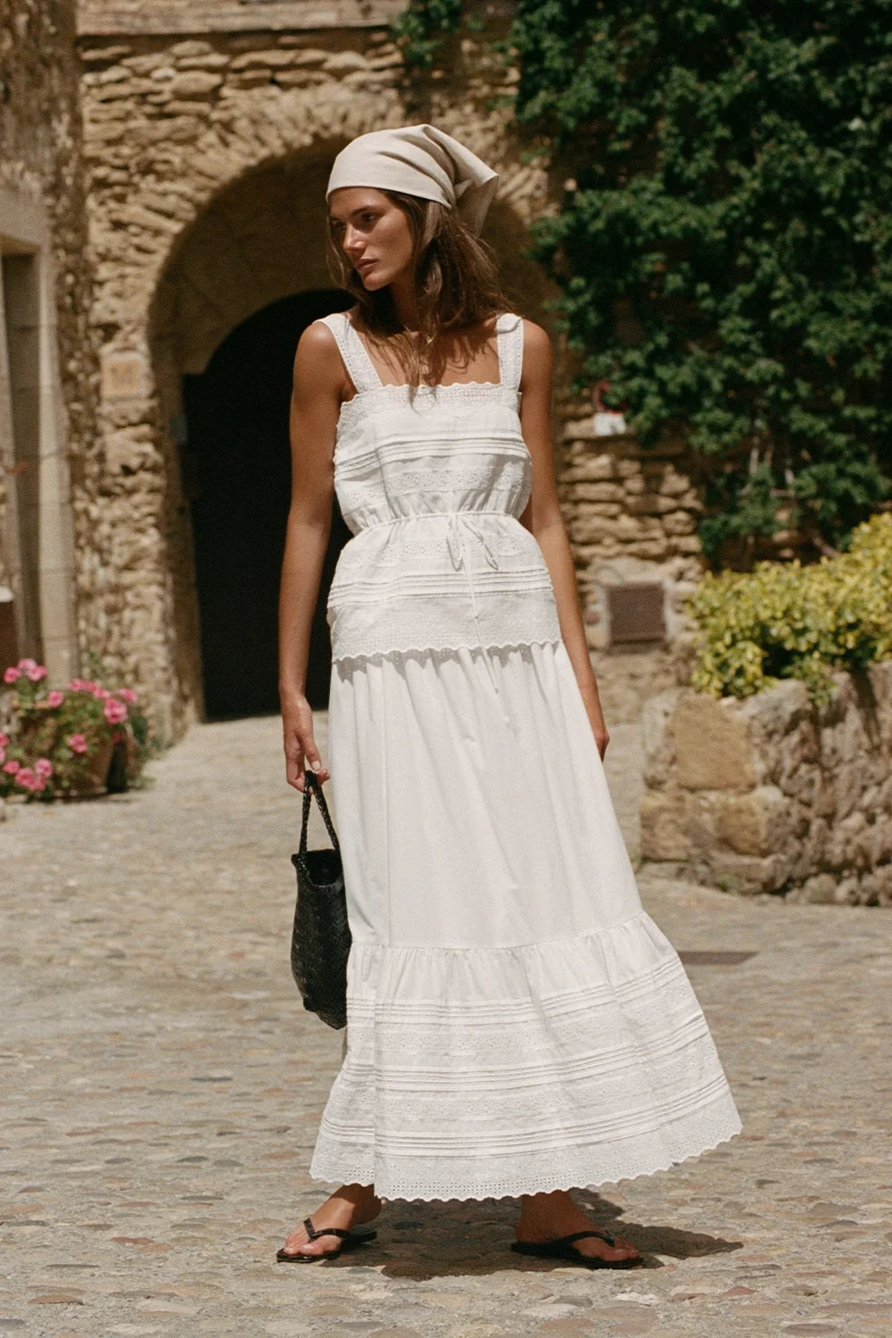 A woman wearing a white tiered maxi dress with a matching headscarf stands on a cobblestone path in front of a rustic stone building.