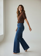 Woman wearing a brown top and blue jeans against a neutral background