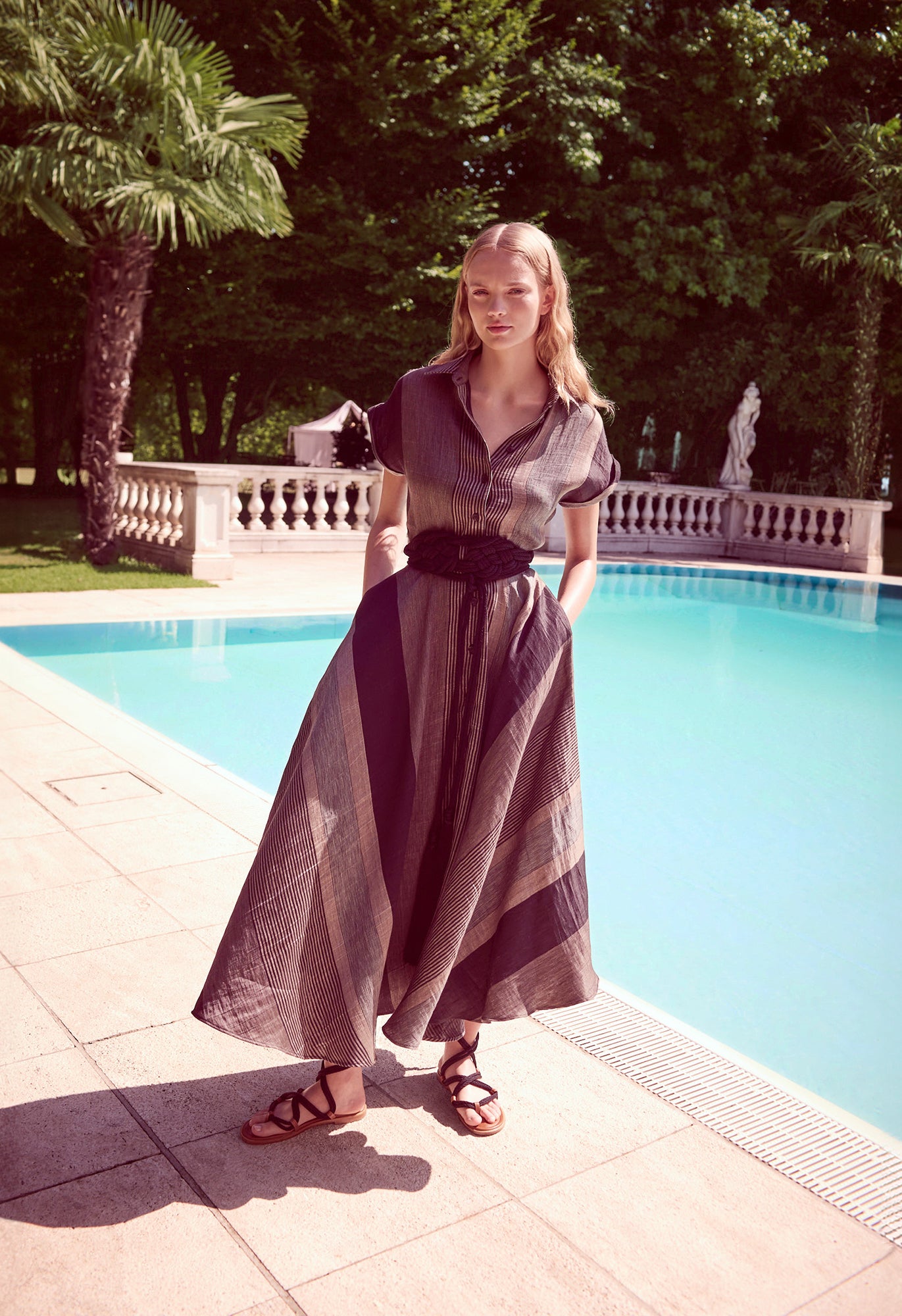 Woman in a long SARA ROKA dress standing by a pool with palm trees in the background.SHOP ONLINE