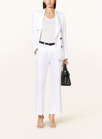 A woman standing, wearing a white linen blazer with a collar, double-breasted button details, and a black belt. She also has a black purse and is wearing a white shirt underneath.