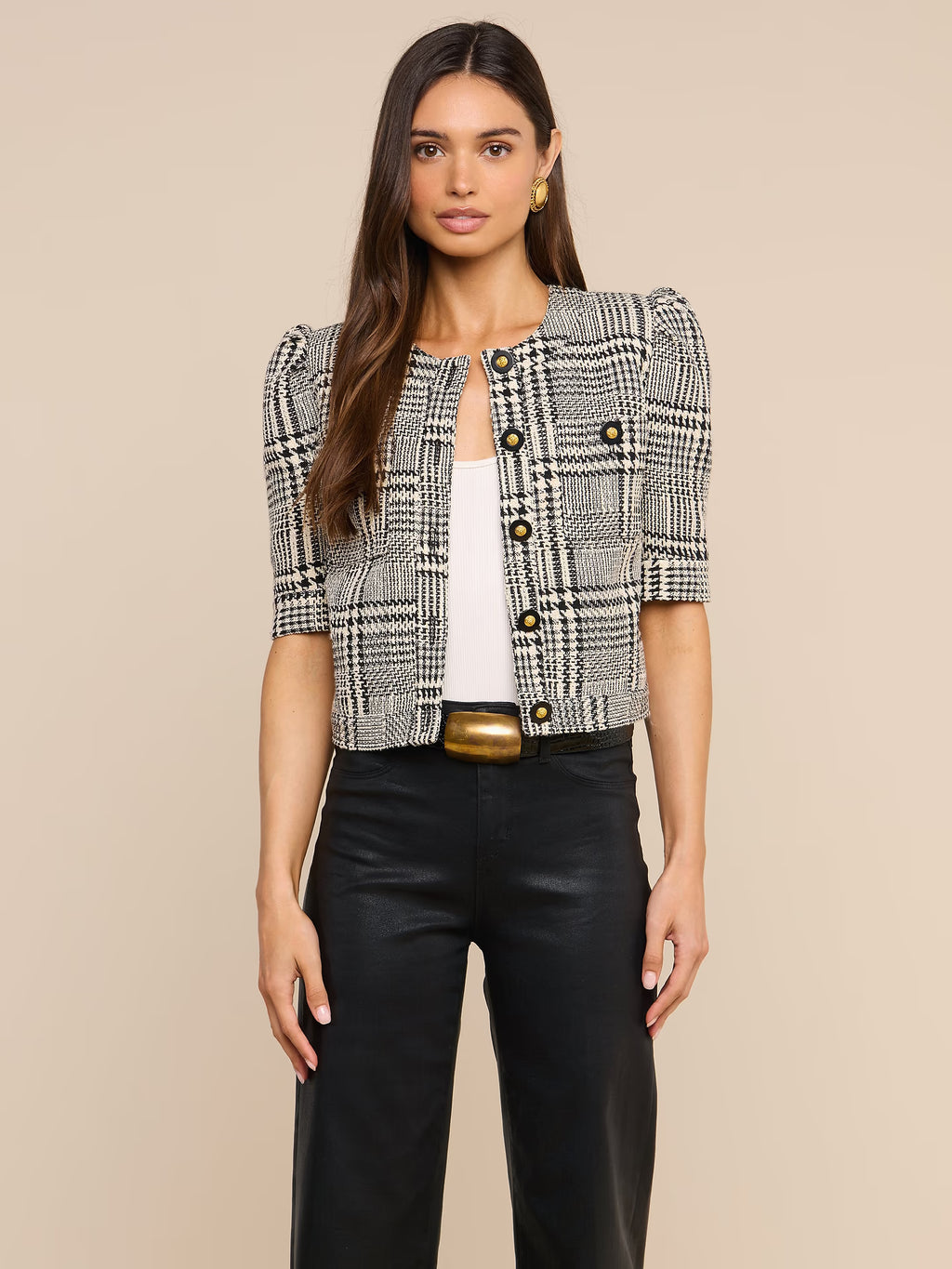 A woman modeling a cropped black and white plaid tweed jacket with puff sleeves and gold buttons, paired with black pants and a gold belt, against a solid beige background.