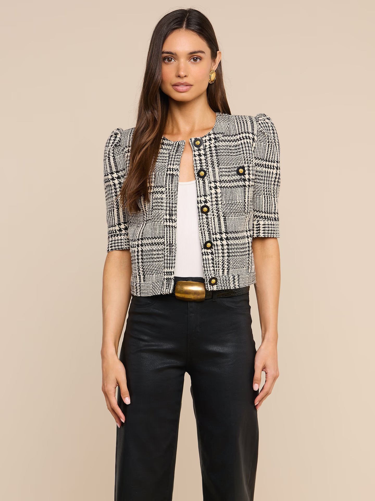 A woman modeling a cropped black and white plaid tweed jacket with puff sleeves and gold buttons, paired with black pants and a gold belt, against a solid beige background.