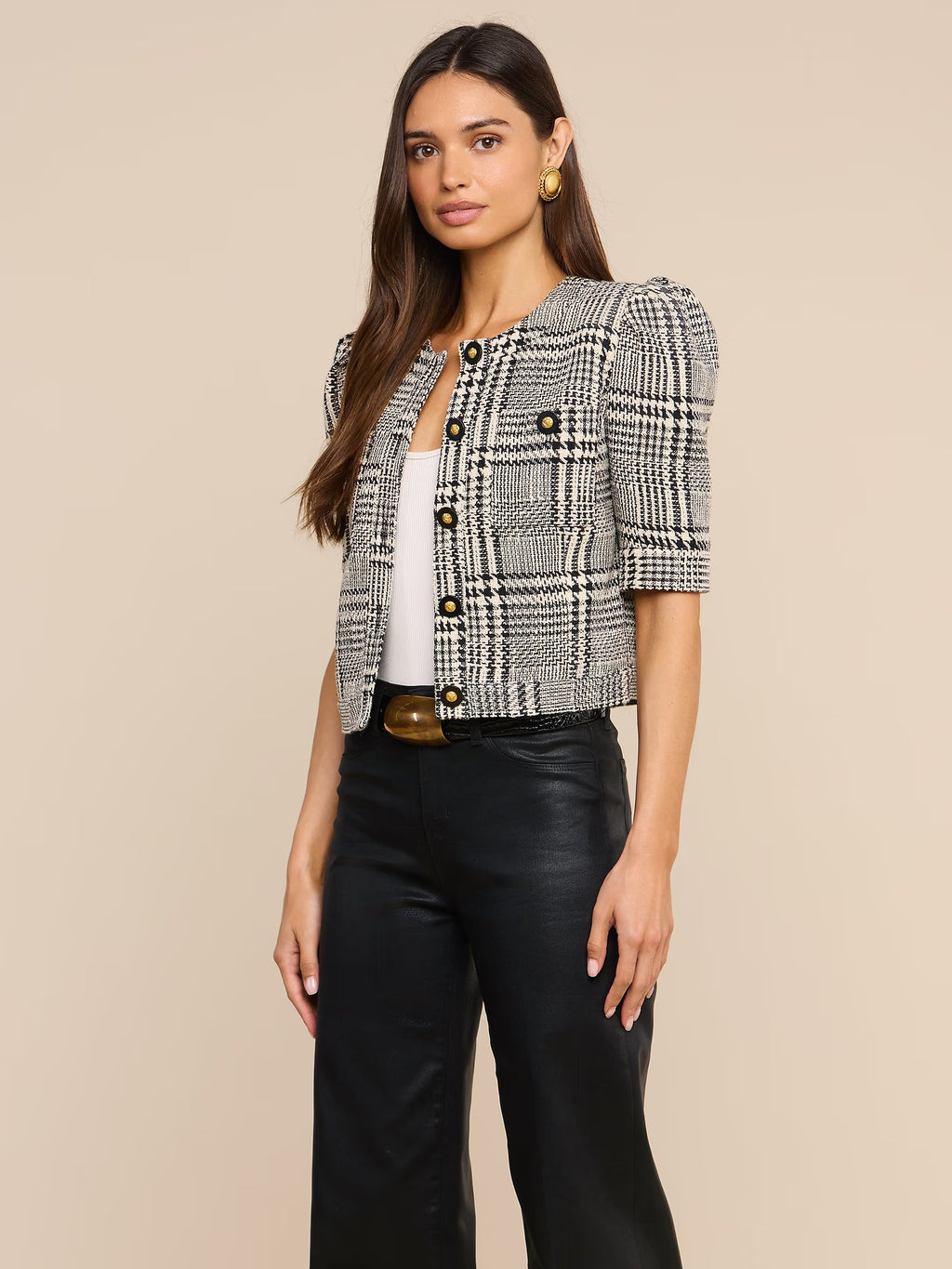 A woman models a short puff-sleeve tweed jacket in a black and white houndstooth pattern, styled with a white top, black leather pants, and a gold belt, against a solid beige background.