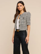 A woman models a short puff-sleeve tweed jacket in a black and white houndstooth pattern, styled with a white top, black leather pants, and a gold belt, against a solid beige background.