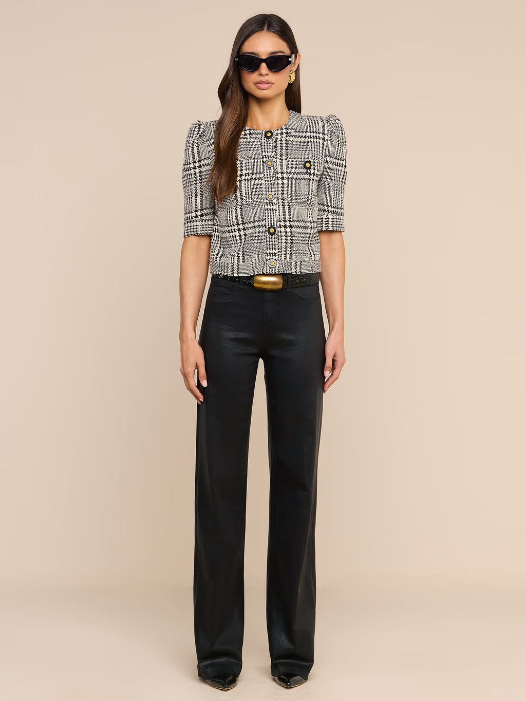 A model stands against a solid beige studio background wearing a black and white plaid cropped jacket with puff sleeves, black wide-leg trousers, a black belt with a gold buckle, and black sunglasses.