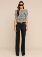 A model stands against a solid beige studio background wearing a black and white plaid cropped jacket with puff sleeves, black wide-leg trousers, a black belt with a gold buckle, and black sunglasses.