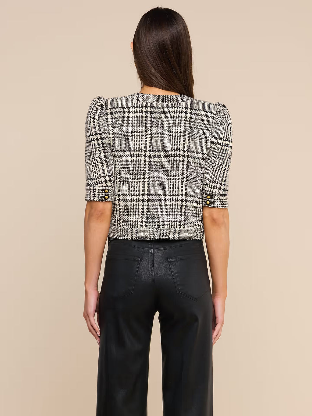 A woman viewed from the back wearing a cropped plaid jacket with puff sleeves and black wide-leg trousers, against a plain beige studio background.