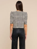 A woman viewed from the back wearing a cropped plaid jacket with puff sleeves and black wide-leg trousers, against a plain beige studio background.
