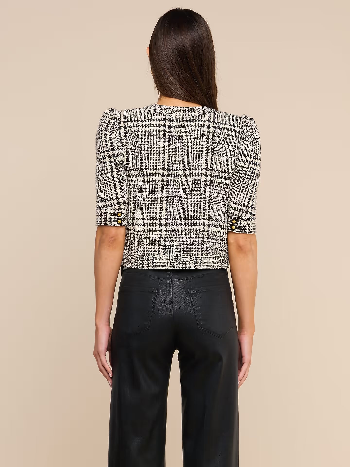 A woman viewed from the back wearing a cropped plaid jacket with puff sleeves and black wide-leg trousers, against a plain beige studio background.