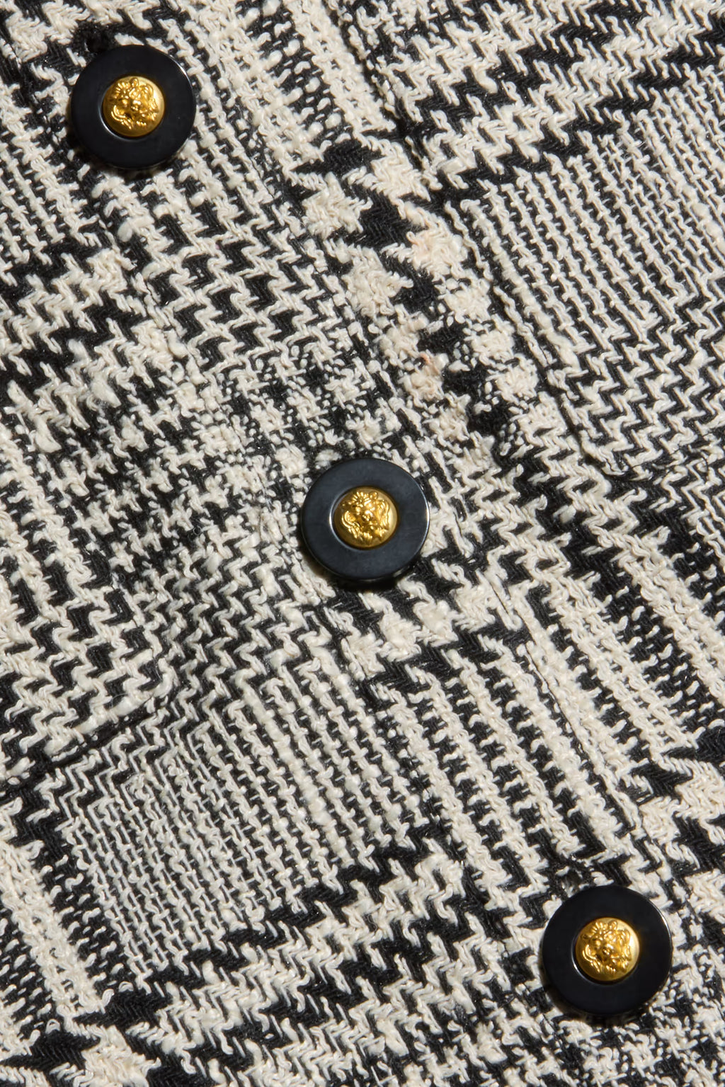 Close-up of a black and off-white woven tweed fabric with a geometric houndstooth-like pattern and decorative round buttons with gold centers.
