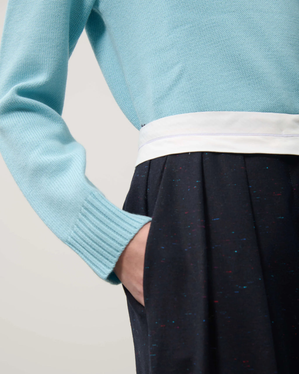 Person wearing a light blue sweater over a white shirt with dark pants on a neutral background