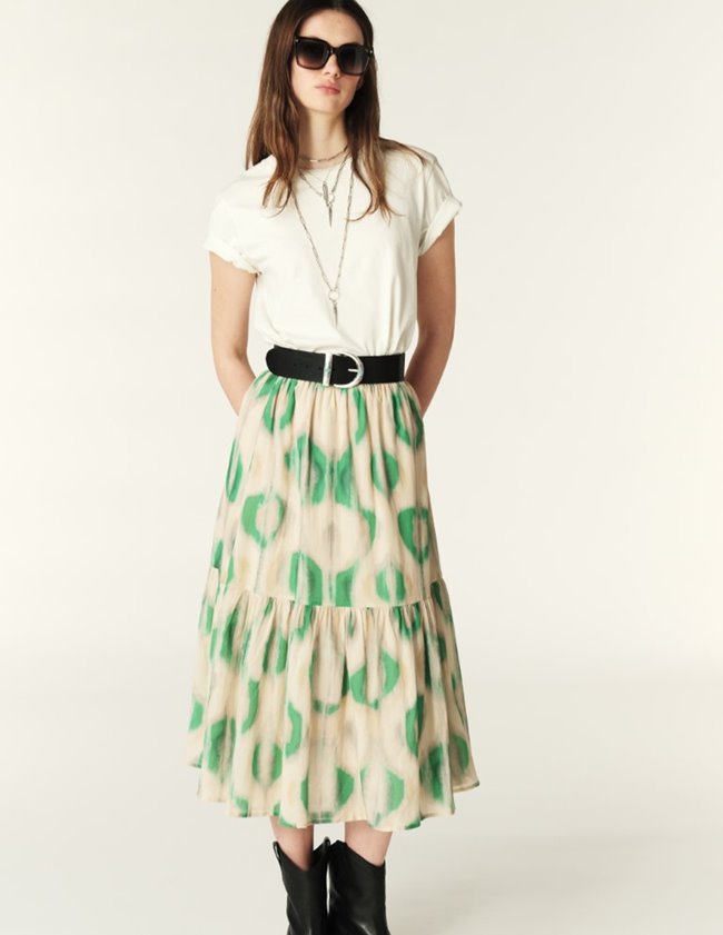 A woman wearing a white top and sunglasses, standing against a plain background. She is also wearing a green and beige floral skirt with a long pleat and an elastic waistband.