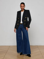 Woman wearing a black blazer with gold buttons and blue jeans on a plain background