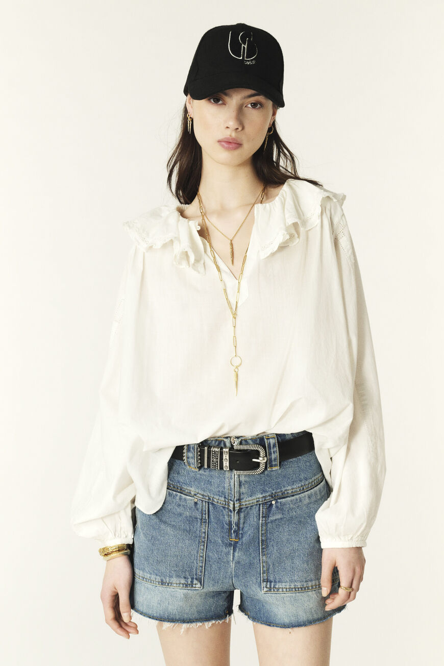 A female model wearing a white loose-fitting blouse with ruffled collar and gathered shoulders, paired with blue denim shorts and a black cap.