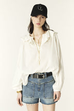 A female model wearing a white loose-fitting blouse with ruffled collar and gathered shoulders, paired with blue denim shorts and a black cap.
