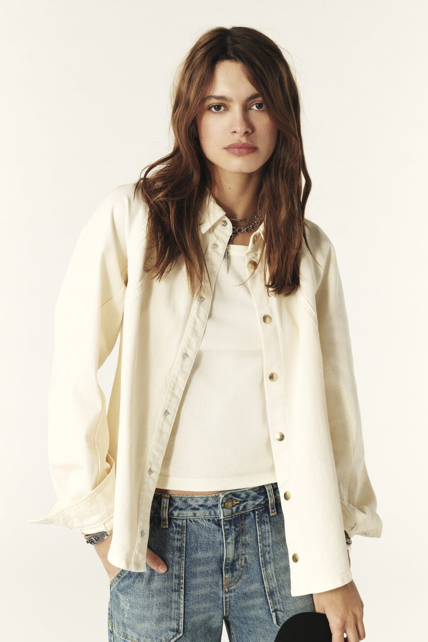 A female model wearing a white denim overshirt with press studs on the front and raglan sleeves. She is paired with blue jeans.