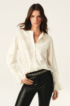 A woman wearing a white long-sleeved blouse with floral embroidery and a V-neck collar, paired with black pants.