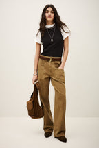 Woman wearing a black top, white shirt, brown pants, and holding a brown bag on a white background