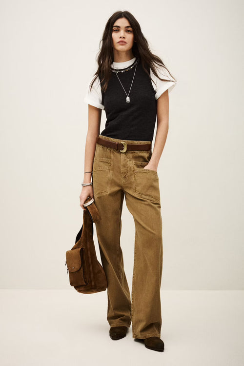 Woman wearing a black top, white shirt, brown pants, and holding a brown bag on a white background