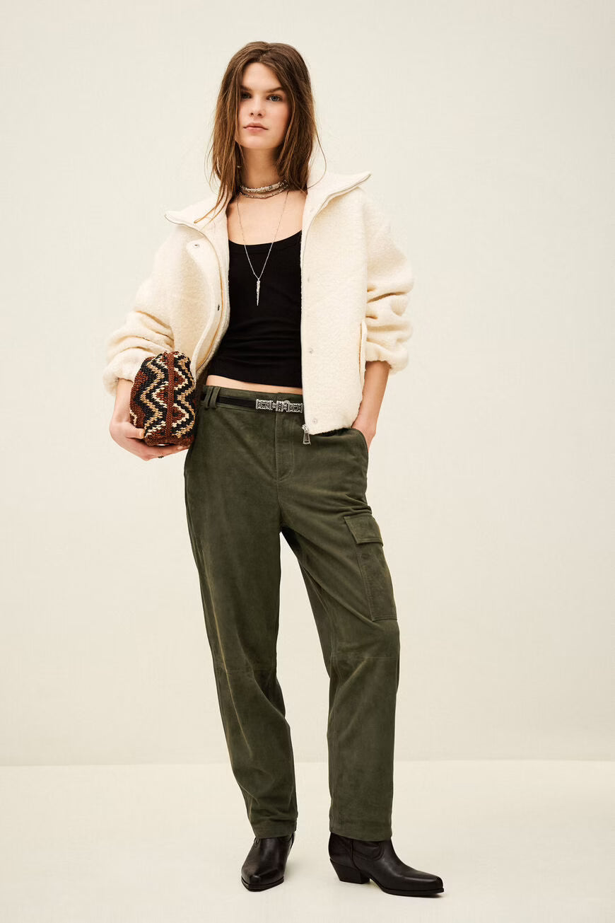 Woman wearing a cream jacket, black top, and green pants holding a patterned clutch against a white background