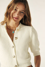 Woman wearing a white textured cardigan with gold buttons on a plain background