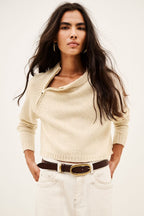 A woman modeling a cream-colored knit sweater with a half-zip neckline, paired with white jeans and a brown belt, against a plain off-white background.
