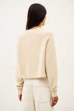 A woman viewed from the back wearing a cropped, long-sleeve knit sweater in a warm cream or oatmeal color, paired with light cream pants, against a plain off-white studio background.