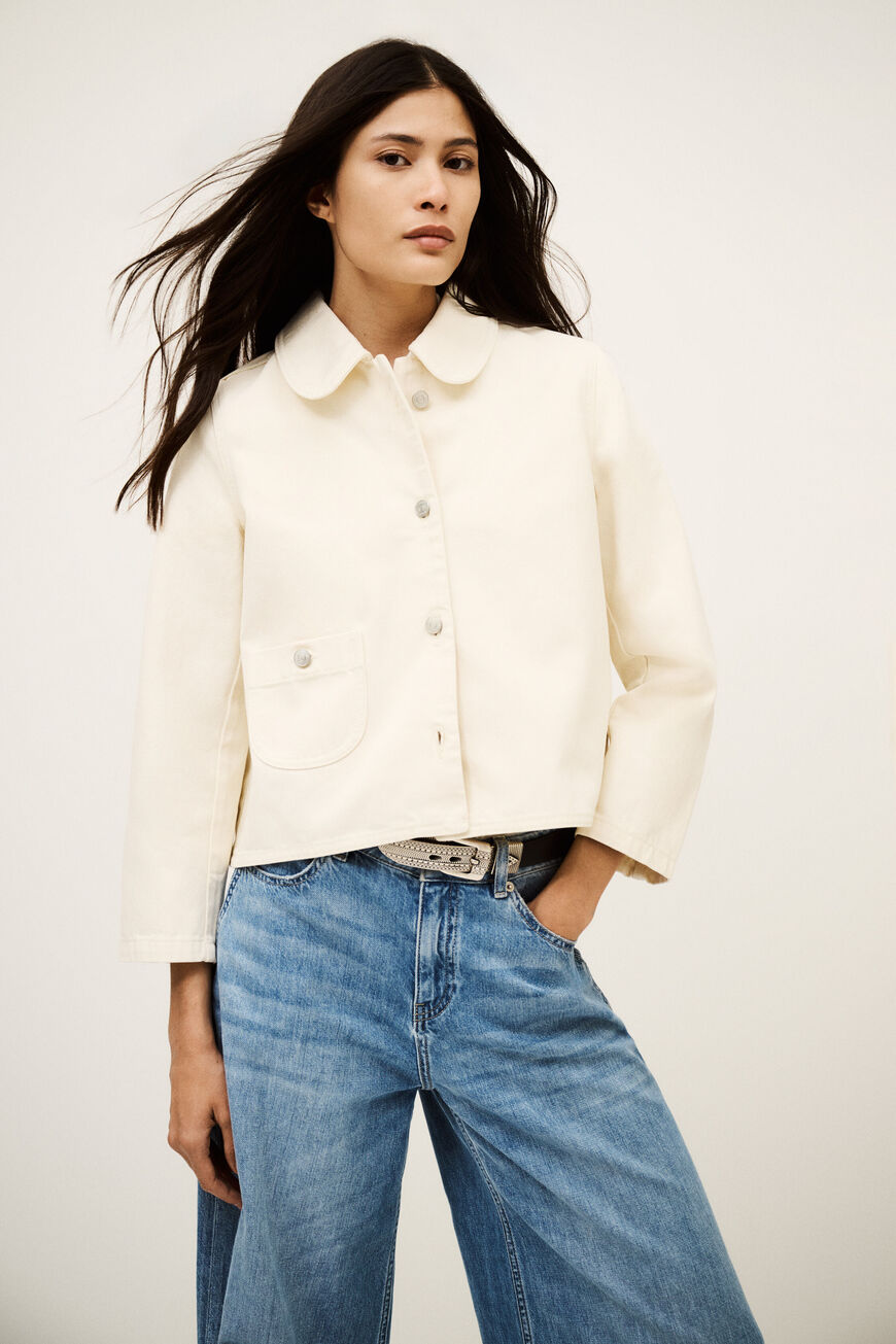 Woman wearing a white jacket and blue jeans against a plain background