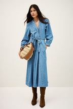 Woman wearing a blue denim dress holding a woven bag on a white background