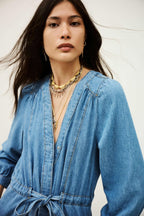 Woman wearing a blue denim dress with layered necklaces against a neutral background