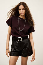 Woman wearing a dark purple top and black leather shorts with a belt on a plain background