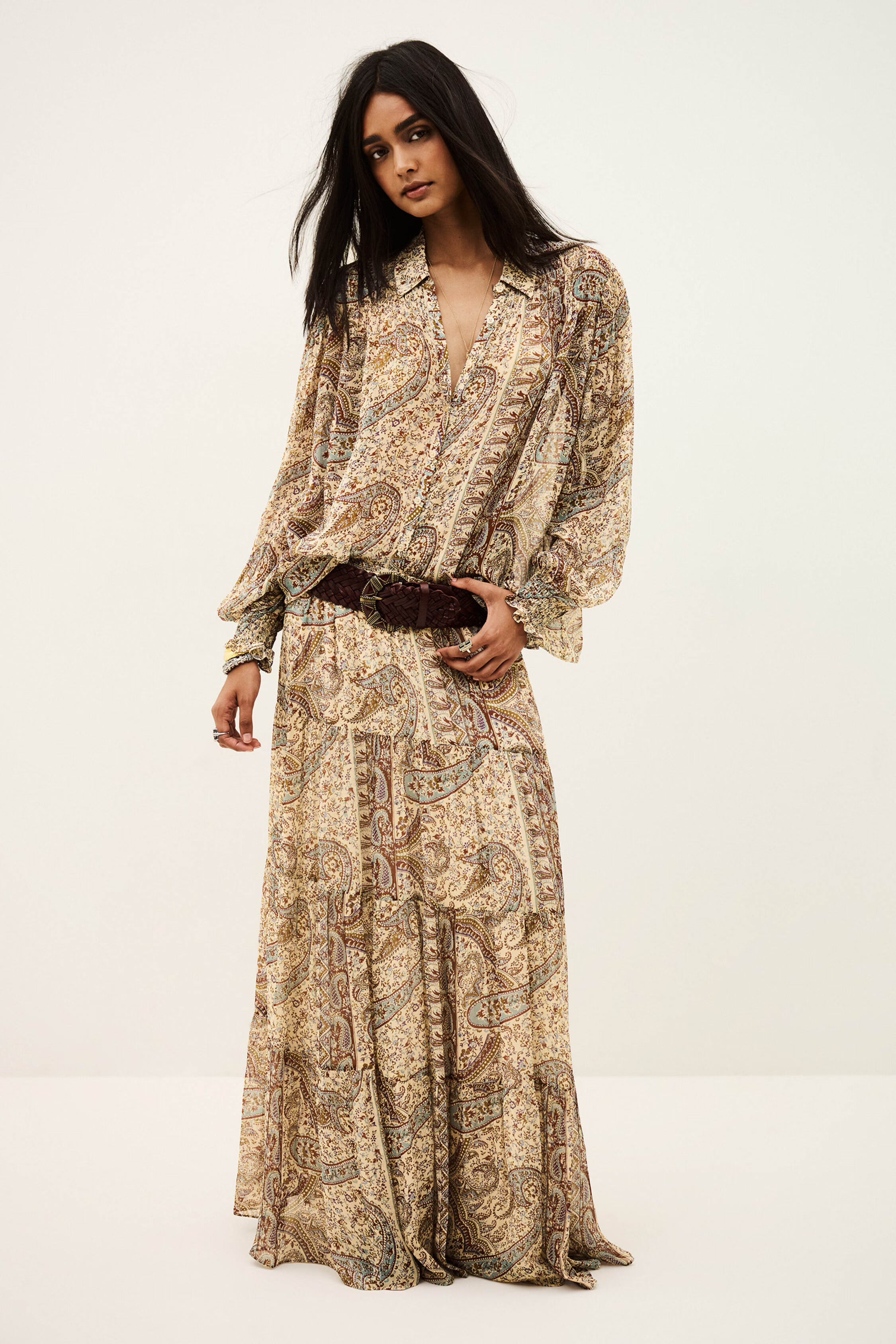 A woman modeling a long-sleeved maxi dress with a beige, brown, and blue paisley pattern, styled with a dark brown textured belt, against a plain off-white studio background.