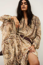 A woman wearing a long-sleeved, loose-fitting bohemian paisley dress in beige, brown, and muted blue, styled with a dark brown braided belt. She is posed against a plain off-white background.
