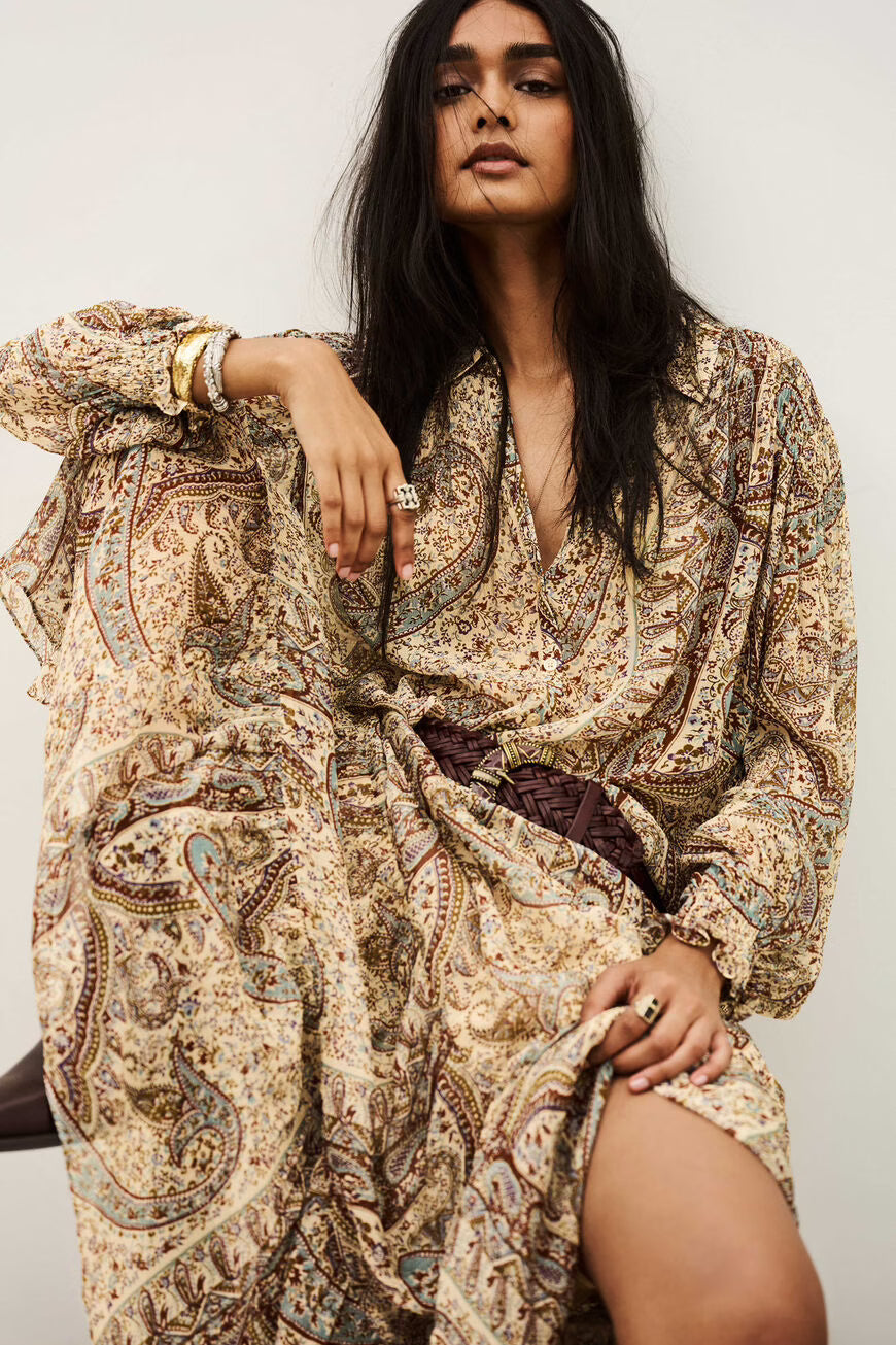 A woman wearing a long-sleeved, loose-fitting bohemian paisley dress in beige, brown, and muted blue, styled with a dark brown braided belt. She is posed against a plain off-white background.