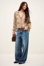 A woman modeling a bohemian-style outfit featuring a beige and brown paisley print blouse, wide-leg blue jeans, a gold-tone belt, and a small brown handbag, set against a plain off-white studio background.
