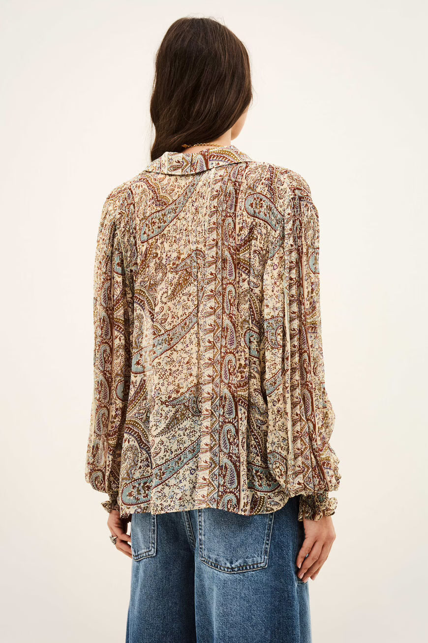 A woman viewed from the back wearing a long-sleeve button-up blouse with an intricate paisley pattern in beige, brown, and red tones, paired with blue jeans against a plain off-white background.