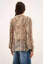 A woman viewed from the back wearing a long-sleeve button-up blouse with an intricate paisley pattern in beige, brown, and red tones, paired with blue jeans against a plain off-white background.