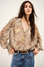 A woman modeling a bohemian paisley print blouse with voluminous sleeves, paired with blue jeans and a brown belt, against a plain light background.