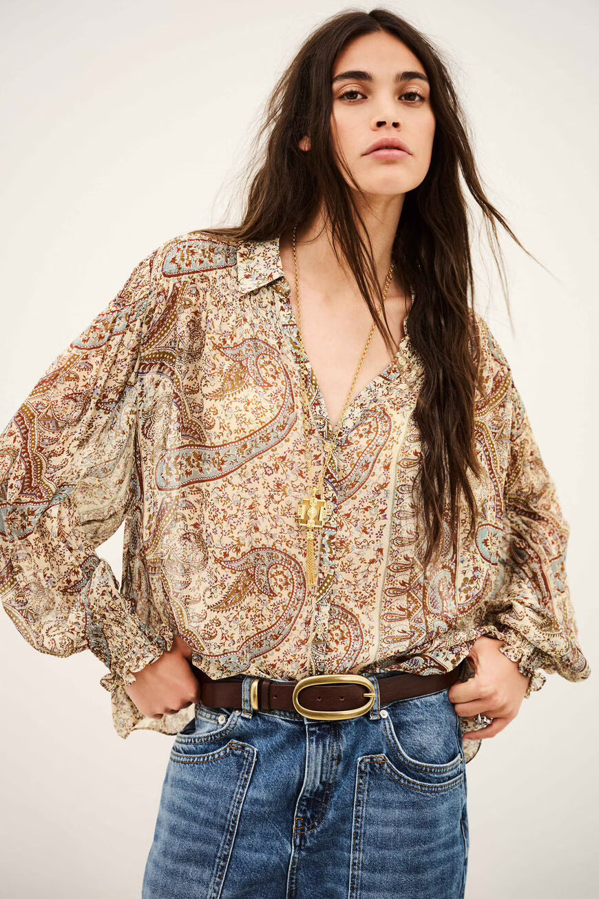 A woman modeling a bohemian paisley print blouse with voluminous sleeves, paired with blue jeans and a brown belt, against a plain light background.