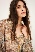 A close-up of a woman wearing a bohemian paisley print blouse with beige, brown, and blue tones, accessorized with a gold chain necklace and ring, against a plain off-white background.