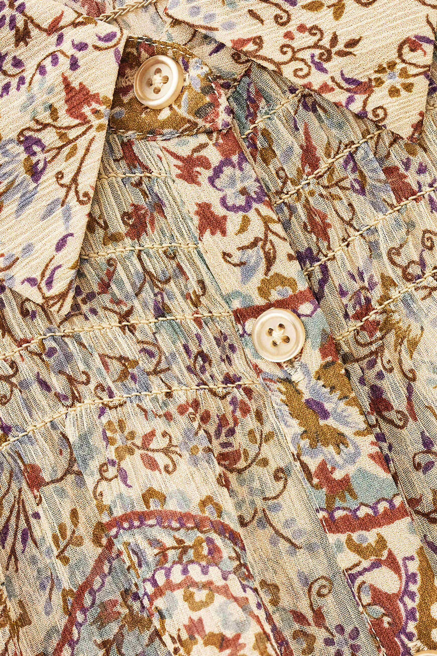 Close-up of a patterned button-up shirt fabric with a cream base and multicolor paisley and floral print in brown, purple, and blue, featuring decorative gold-toned stitching and cream buttons.