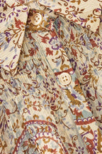 Close-up of a patterned button-up shirt fabric with a cream base and multicolor paisley and floral print in brown, purple, and blue, featuring decorative gold-toned stitching and cream buttons.